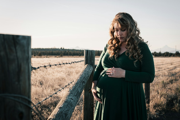 Plus-size pregnant woman in a green wrap dress, cradling her belly with both hands in a field at golden hour