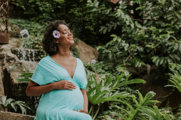 Pregnant woman in a teal V-neck wrap dress smiling with eyes closed in a lush garden setting